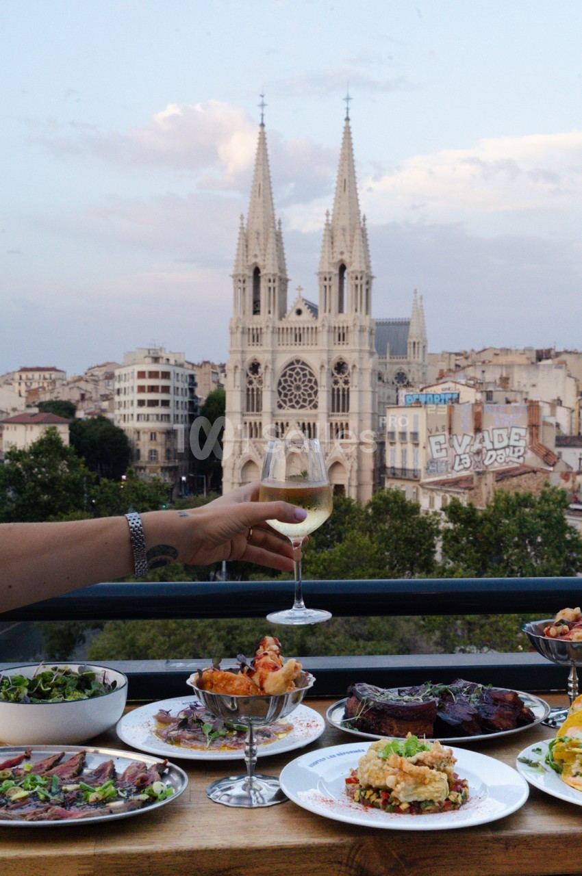 Terrasse avec vue sur la cathédrale de Marseille, table dressée avec plats et verre de vin levé.