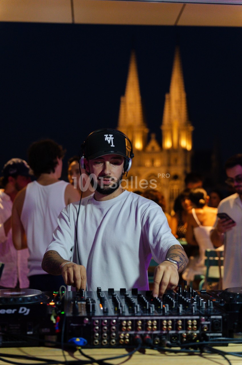 DJ en train de mixer sur platine avec casque, devant une église illuminée en arrière-plan et des convives autour.