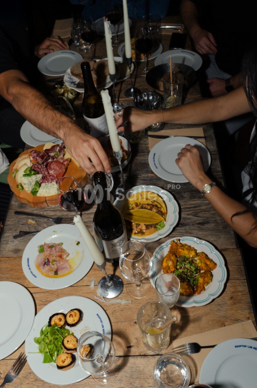 Table en bois dressée avec convives partageant un repas - assiettes de cuisine gastronomique, bouteille de vin et bougies blanches.