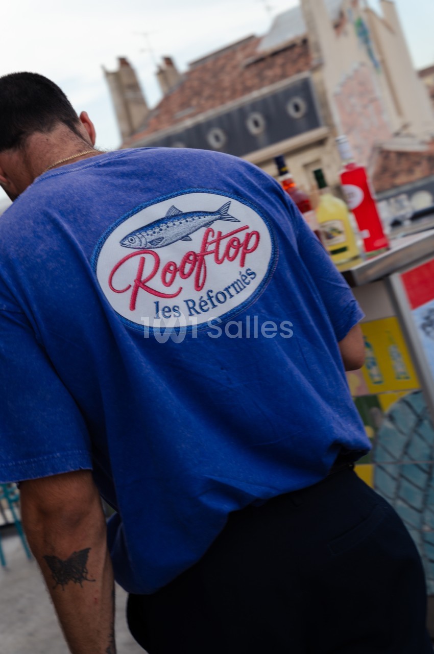 Personne de dos portant un t-shirt bleu avec logo Rooftop les Réformés, place urbaine avec bâtiments anciens.