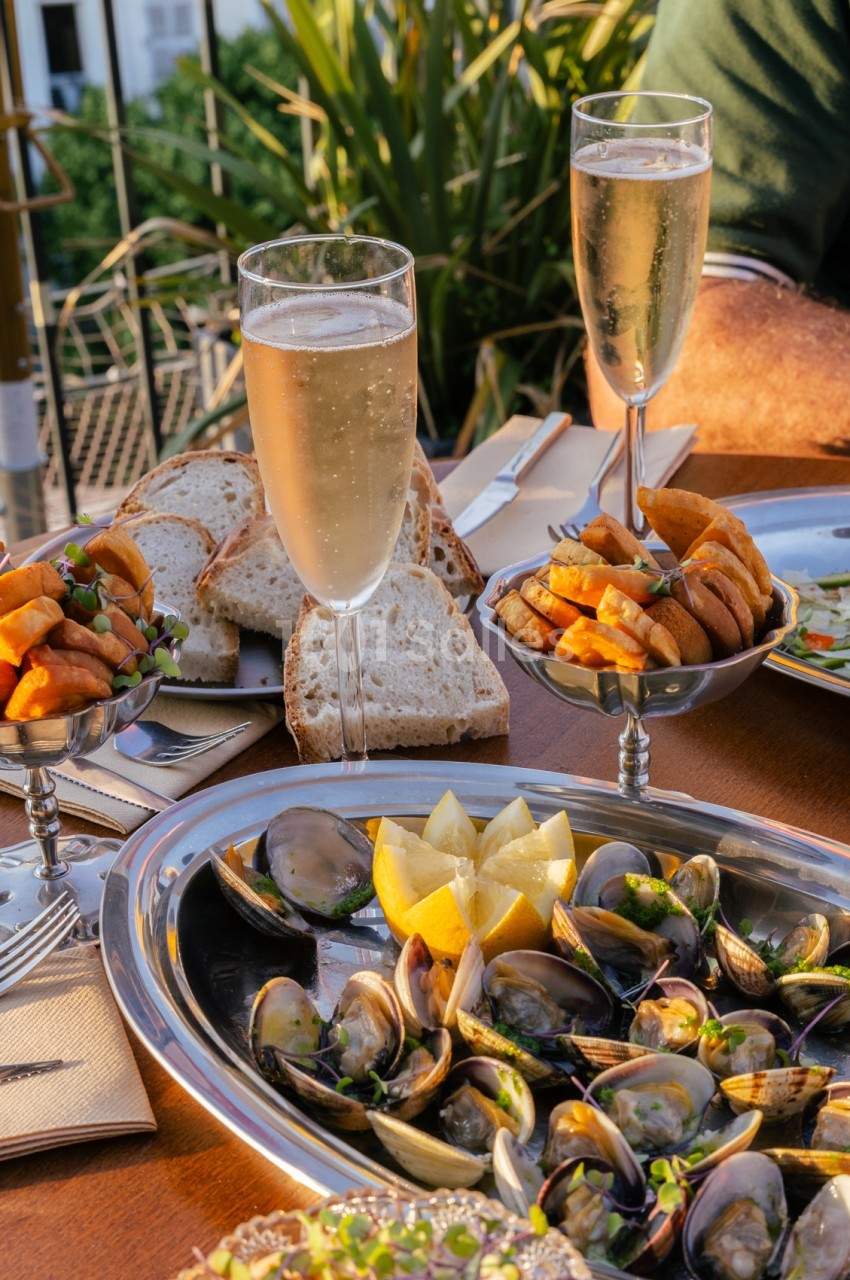 Table dressée en terrasse avec moules aux citrons, pain, verres de champagne et plantes - ciel bleu en arrière-plan.
