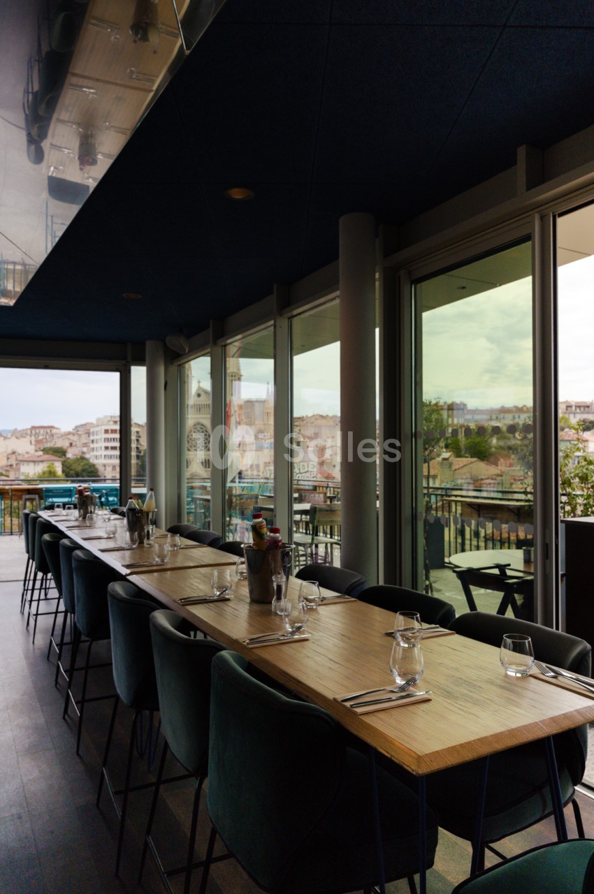 Salle avec tables hautes en bois, chaises noires, baies vitrées ouvertes sur terrasse et vue panoramique urbaine.