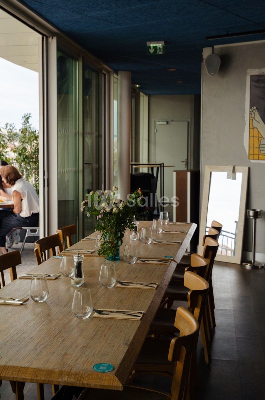 Salle intérieure dressée avec table longue en bois, chaises claires, verres et couverts, baies vitrées ouvertes vers terrasse extérieure.