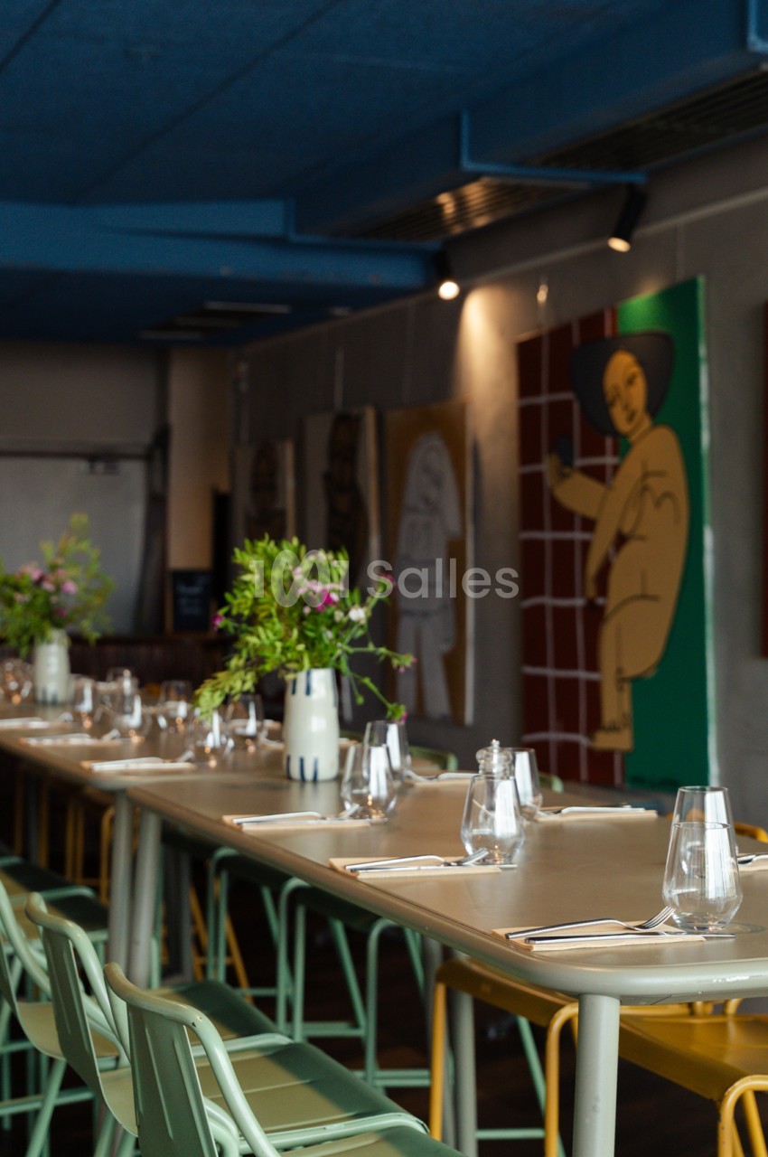Salle à table longue dressée avec verres et couverts, chaises colorées, plantes en vases, murs gris et artwork vert.