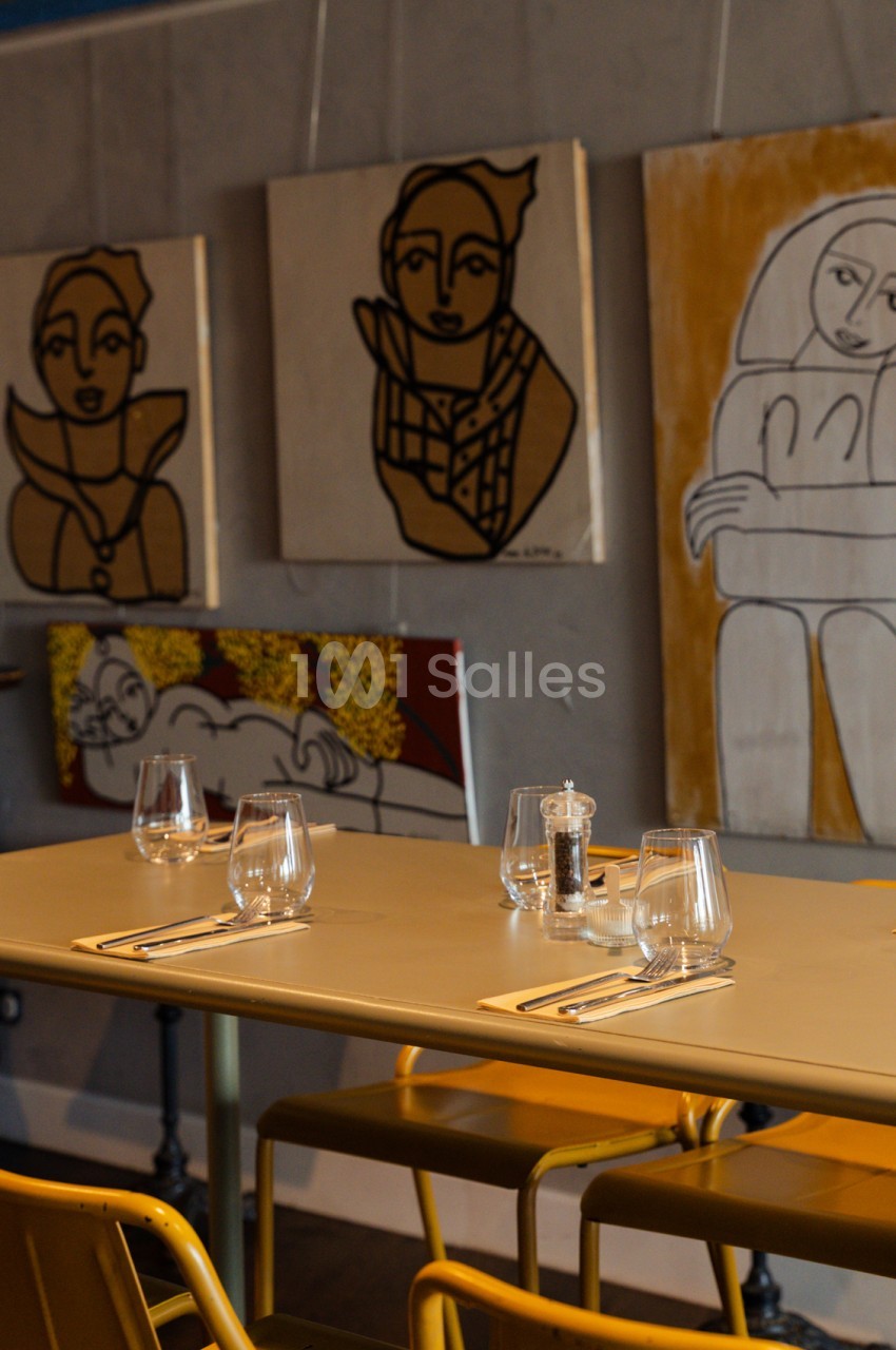 Table dressée avec verres et couverts, chaises jaunes, murs gris ornés de dessins abstraits en noir et ocre.