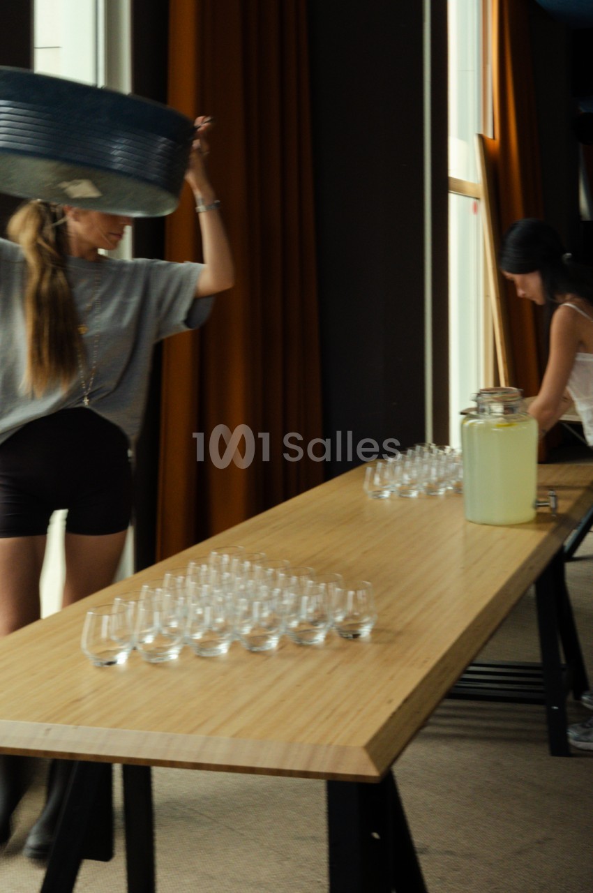 Table en bois dressée avec verres vides et une carafe, deux personnes en arrière-plan devant des rideaux.