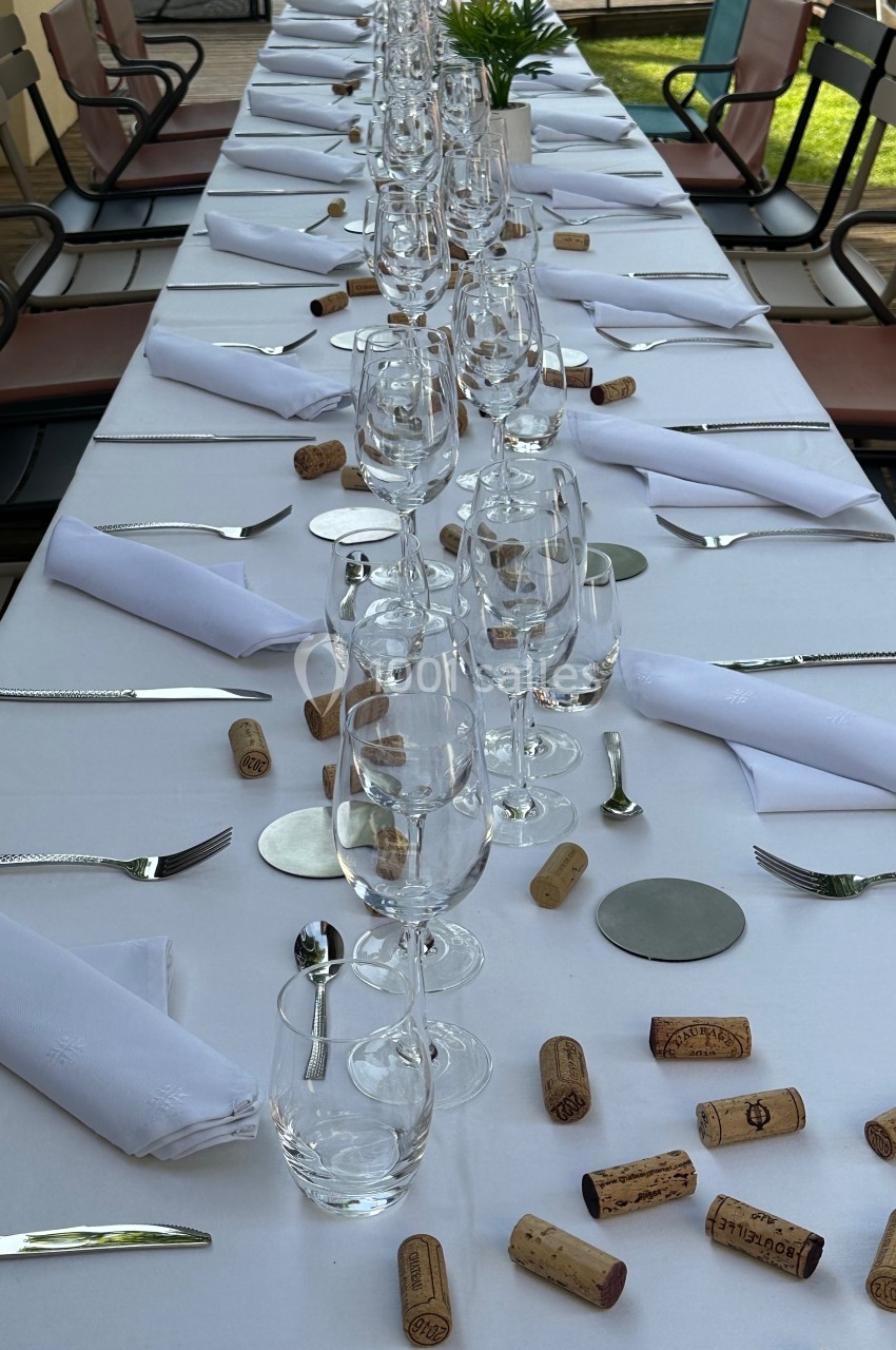 Table dressée avec nappes blanches, verres, couverts, serviettes pliées et bouchons de liège dispersés.
