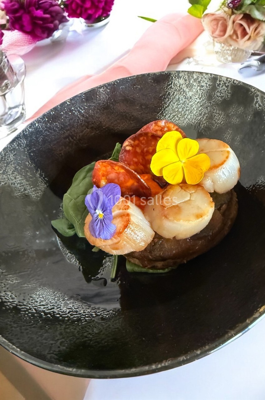 Assiette gastronomique avec noix de Saint-Jacques, tranches de chorizo, fleurs comestibles et lit de légumes verts.