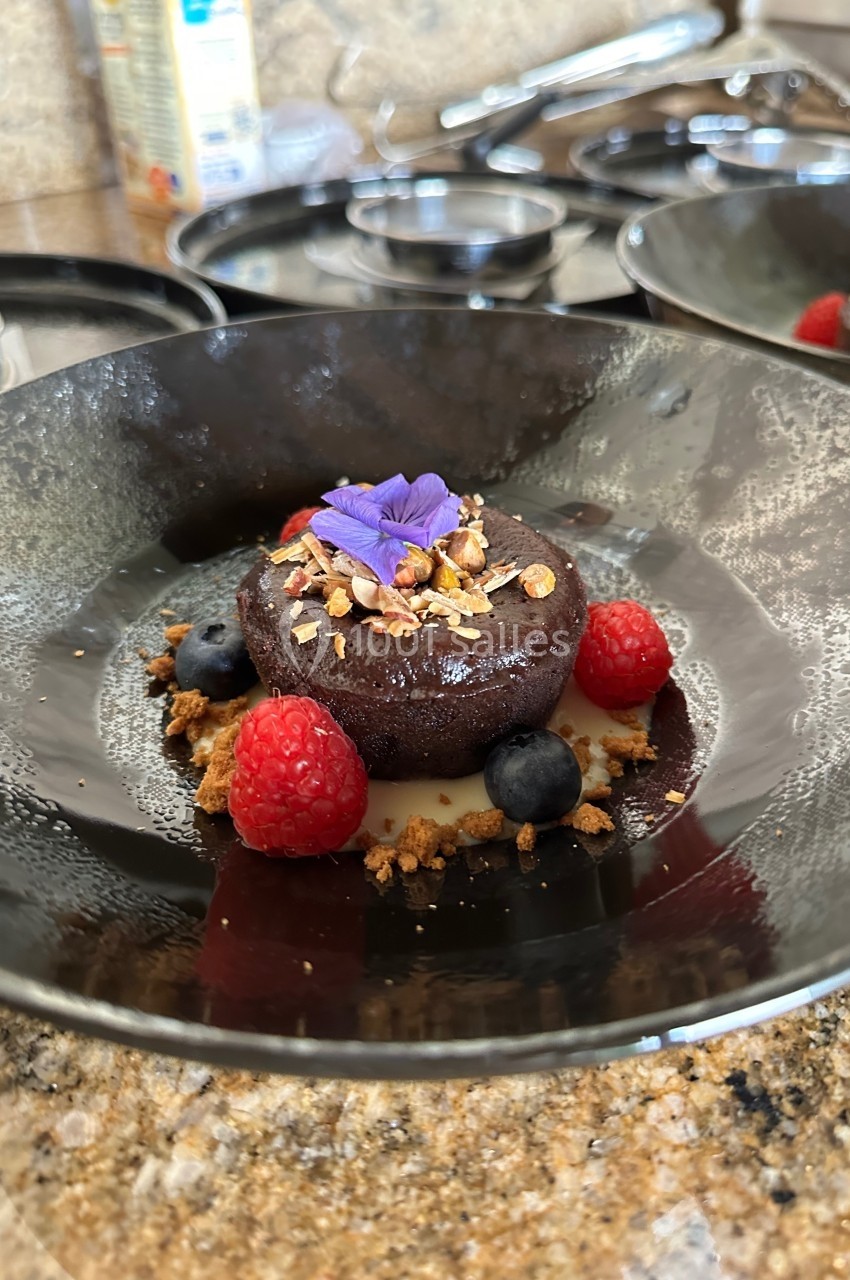 Dessert au chocolat décoré de fruits rouges, myrtilles, granola et une fleur comestible, présenté dans une assiette noire.