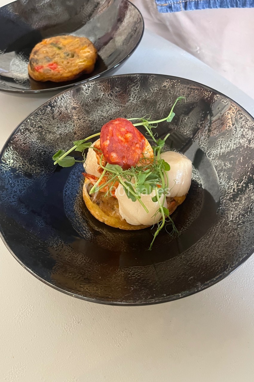Assiette contenant une préparation garnie de légumes, de noix de Saint-Jacques, de chorizo et de pousses vertes.