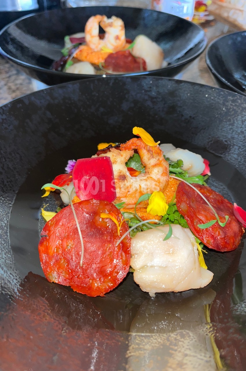 Assiette gastronomique avec crevette, chorizo, légumes colorés et fleurs comestibles dans un bol noir.