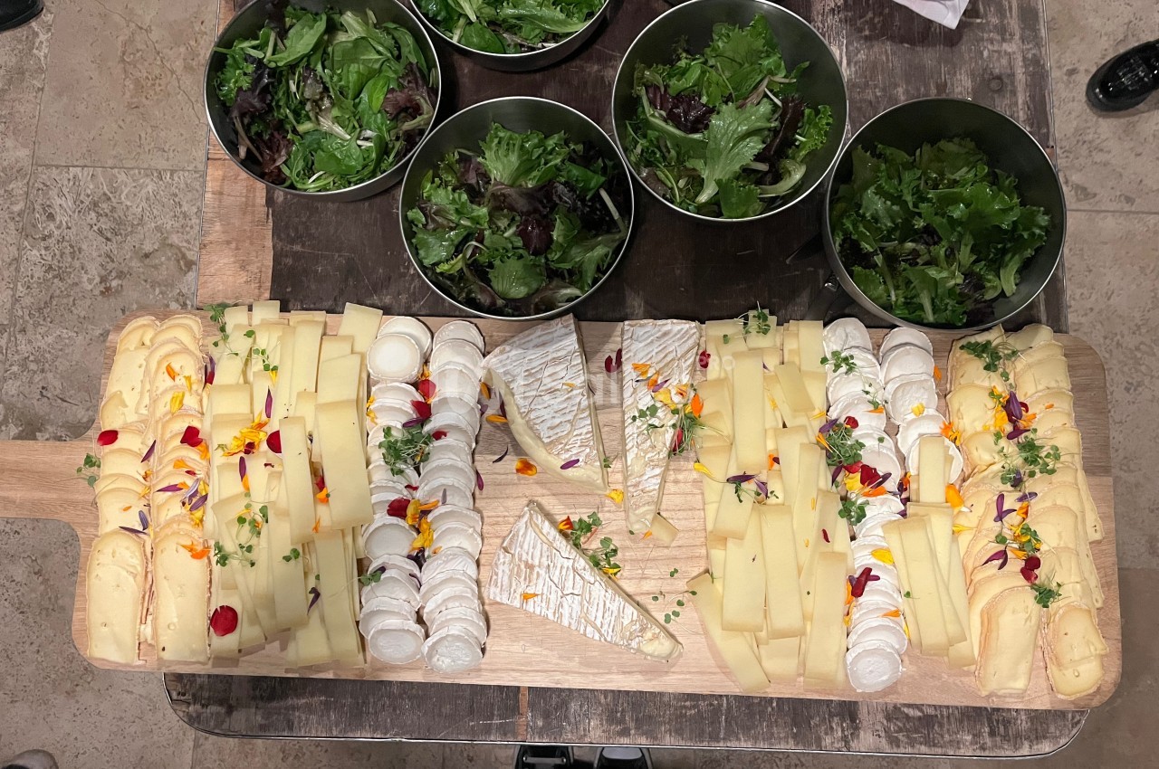 Plateau de fromages variés décoré de fleurs comestibles, accompagné de bols de salade verte sur une table en bois.