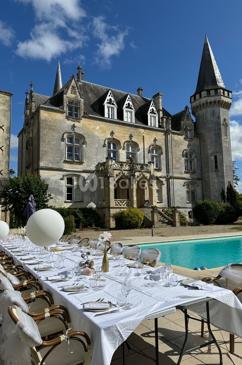 Grande table dressée près d'une piscine, devant un château avec tourelle sous un ciel bleu partiellement nuageux.