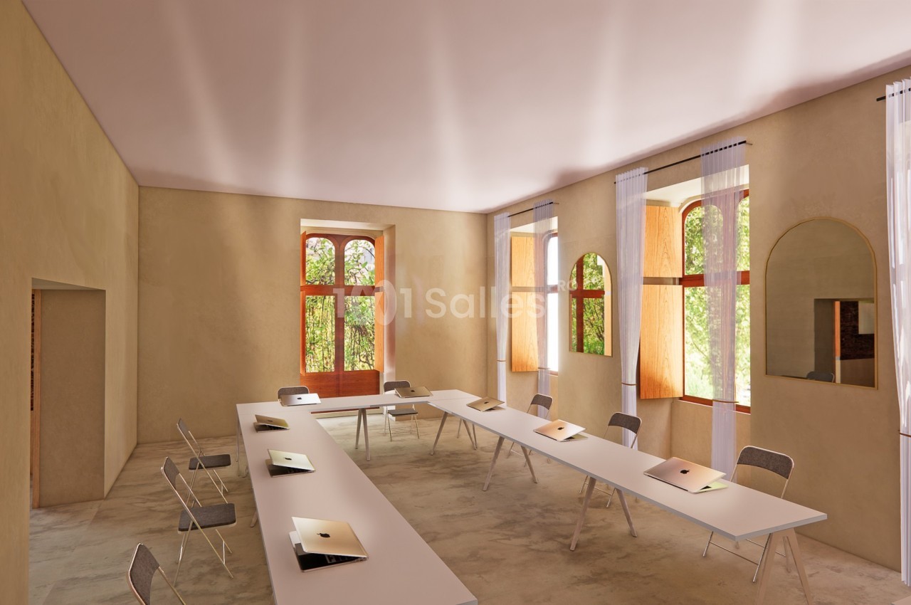Salle lumineuse avec de grandes fenêtres, tables blanches alignées, chaises pliantes et ordinateurs portables posés.