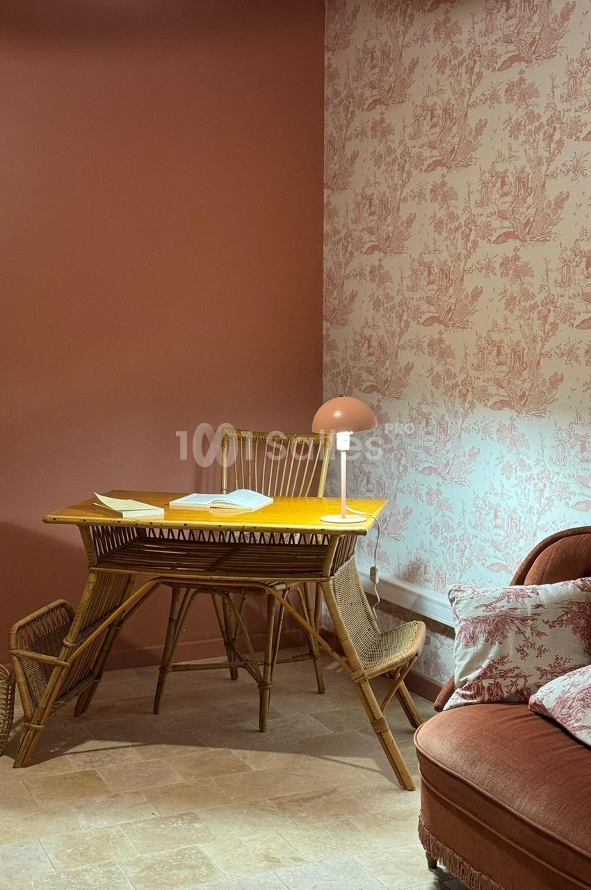 Coin bureau avec table en rotin, lampe allumée, mur rose uni et papier peint à motifs, ambiance chaleureuse.
