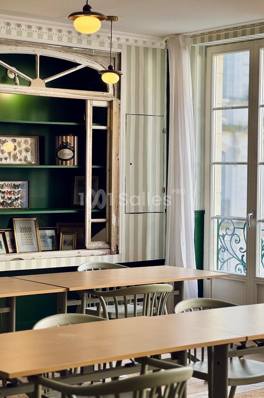 Salle lumineuse avec tables en bois, chaises vertes, étagères décorées et fenêtre donnant sur un balcon en fer forgé.