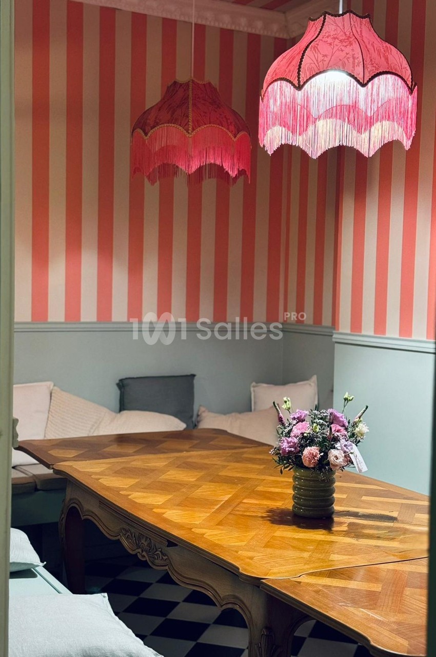Salle à manger avec table en bois, bouquet de fleurs, banquettes et lampes suspendues roses à franges.