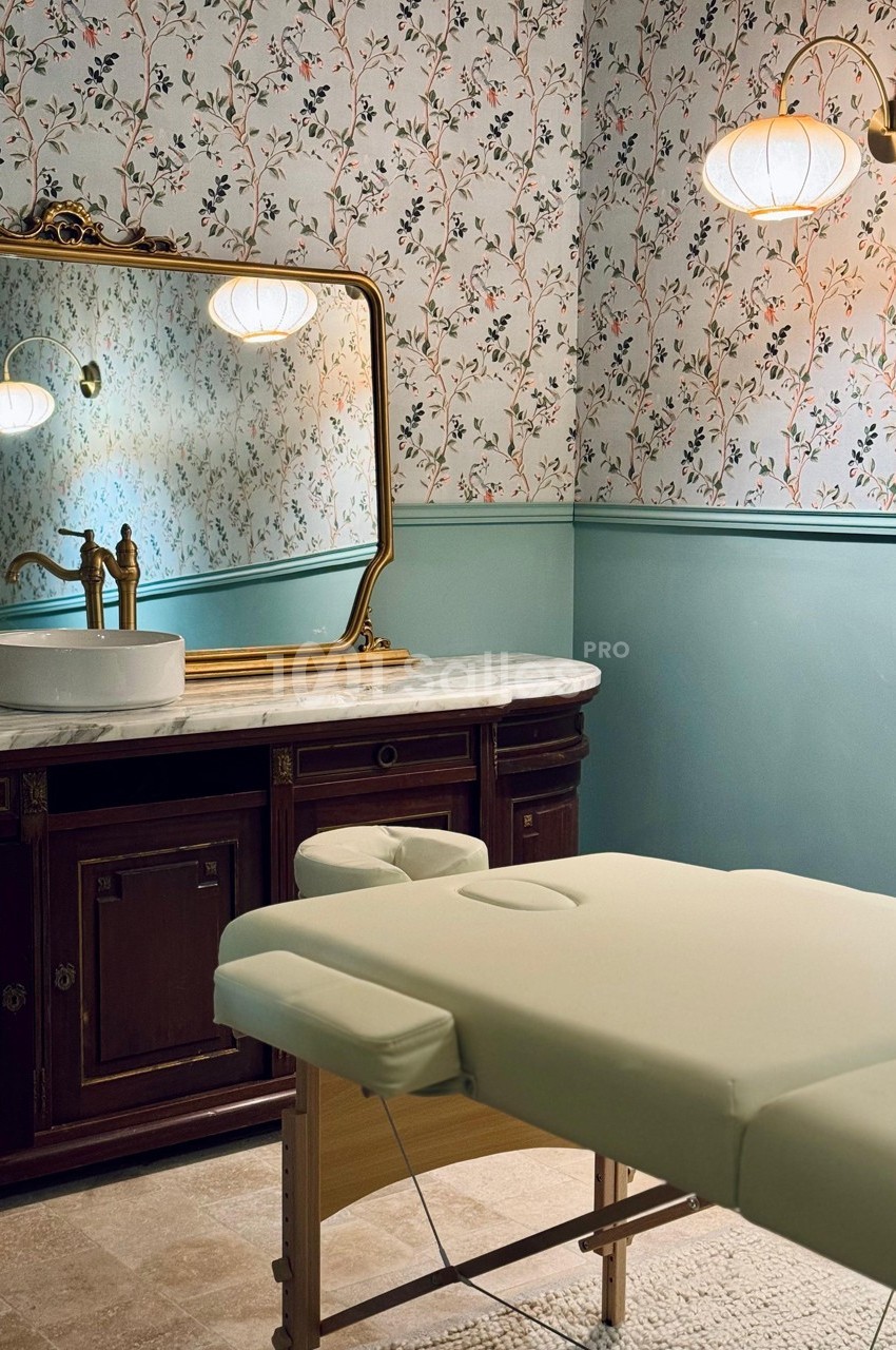 Salle de soin avec table de massage beige, lavabo sur meuble en bois et miroir encadré, éclairée par des appliques murales.