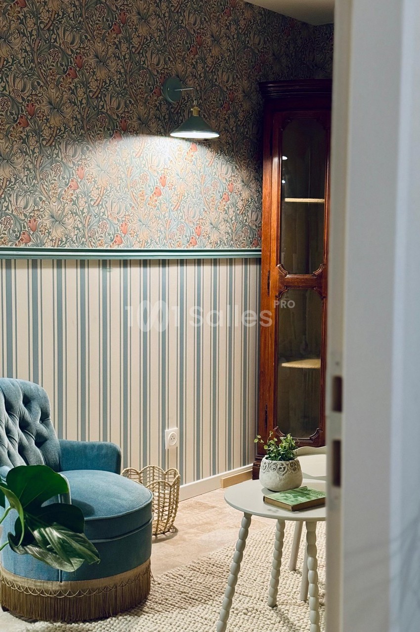 Fauteuil bleu et table basse dans un salon au style vintage avec papier peint fleuri et armoire en bois.
