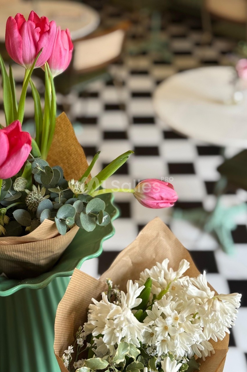 Bouquets de tulipes roses et de fleurs blanches sur un sol en damier noir et blanc, avec des tables floues en arrière-plan.