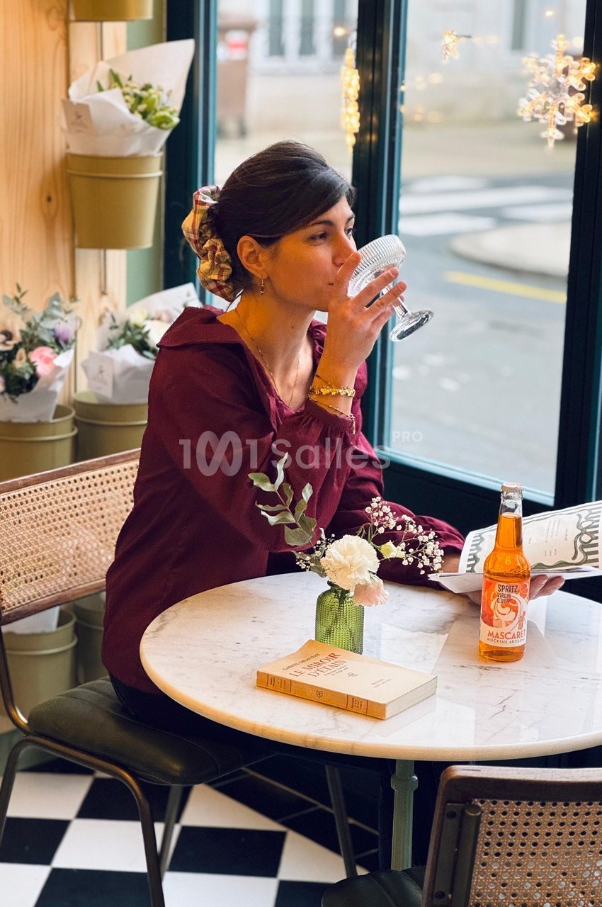 Une femme assise dans un café, buvant dans un verre, avec un livre et une bouteille posés sur une table.