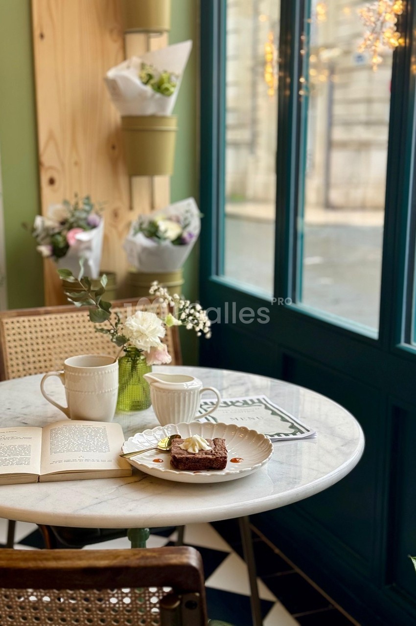 Table ronde en marbre avec deux tasses, un dessert et un livre, près d'une fenêtre lumineuse et de fleurs décoratives.