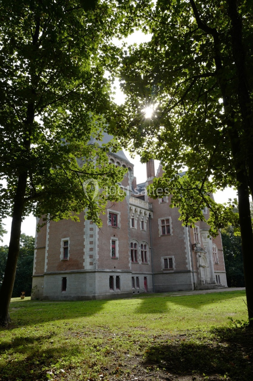 Château en briques entouré d'arbres, avec des rayons de soleil filtrant à travers le feuillage.