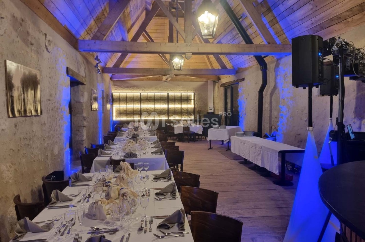 Salle de réception rustique avec poutres apparentes, tables dressées pour un repas et éclairage tamisé bleu.