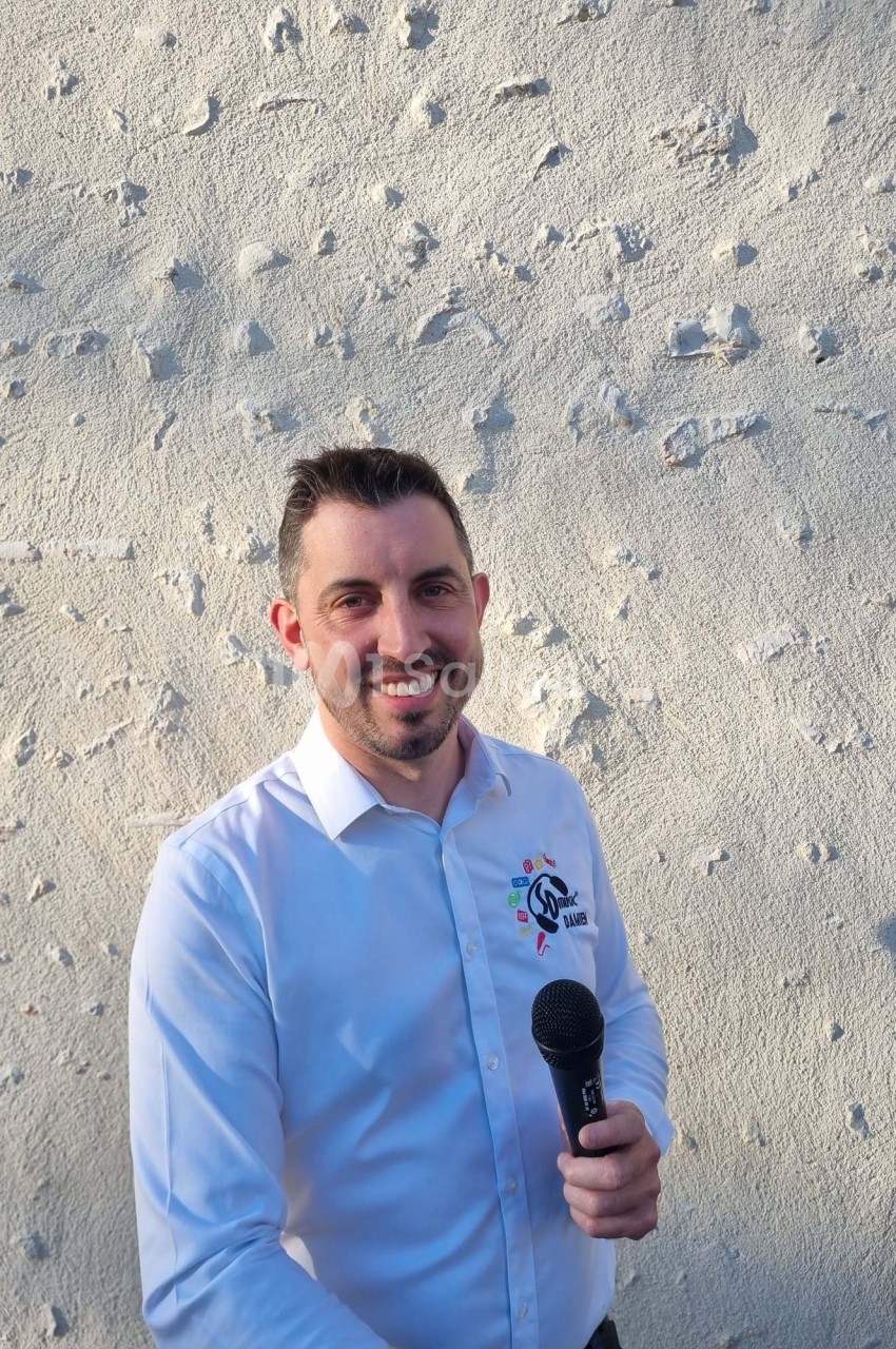 Homme souriant en chemise blanche tenant un micro devant un mur texturé clair.