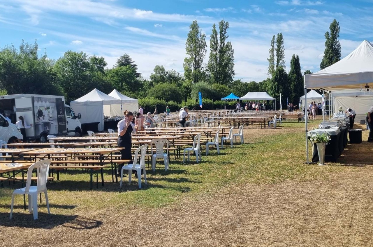 Tables et chaises disposées en extérieur sur une pelouse, avec des stands, des tentes et des camions en arrière-plan.