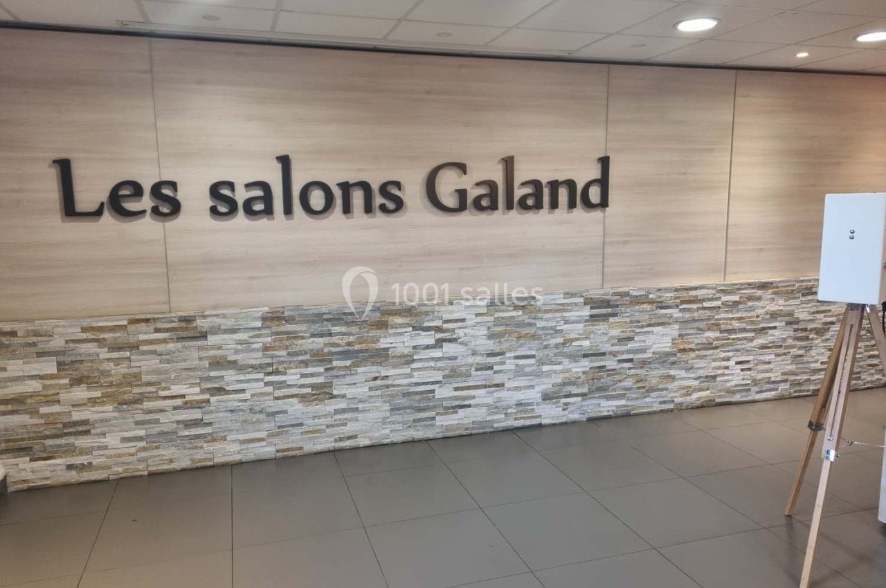 Mur intérieur avec le logo ’Les salons Galand’, décoré de bois clair et de pierres, éclairé par un plafond lumineux.