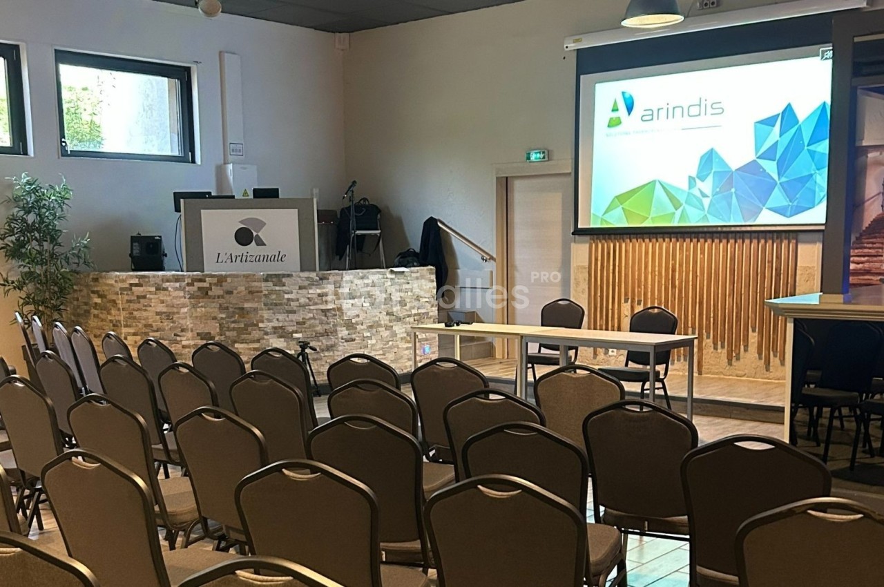 Salle de conférence avec chaises alignées, écran de projection allumé et pupitre sur une estrade en pierre.