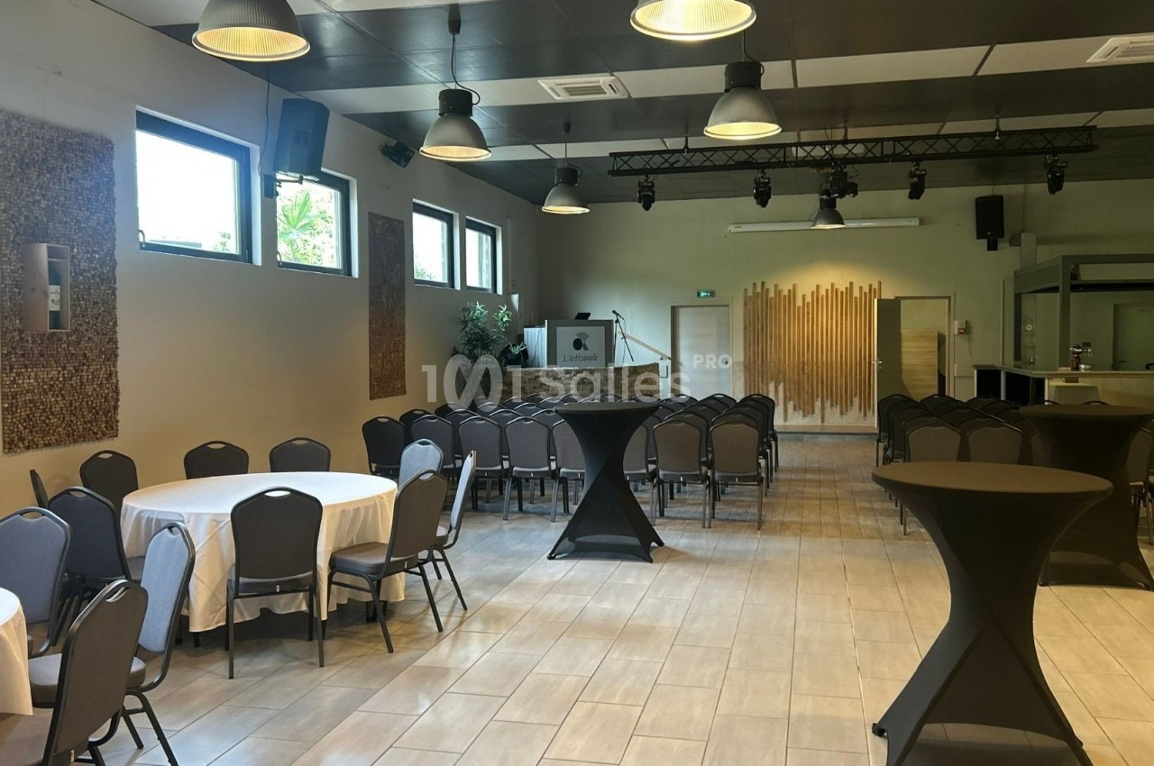 Salle de réception lumineuse avec tables rondes dressées, chaises alignées et un espace scénique en arrière-plan.