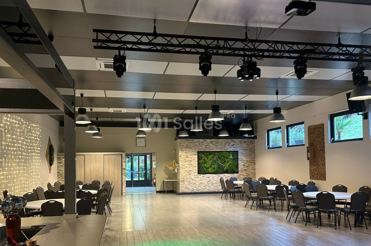 Salle spacieuse avec tables rondes, chaises, éclairage suspendu, murs décorés et grandes fenêtres donnant sur l'extérieur.