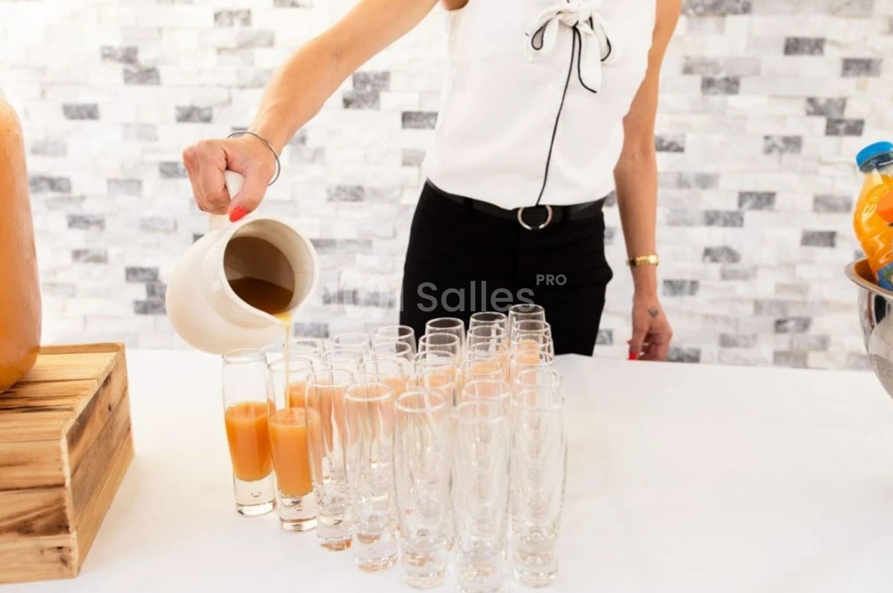 Une personne verse un jus orange dans des verres alignés sur une table blanche, avec un fond de mur en pierre claire.