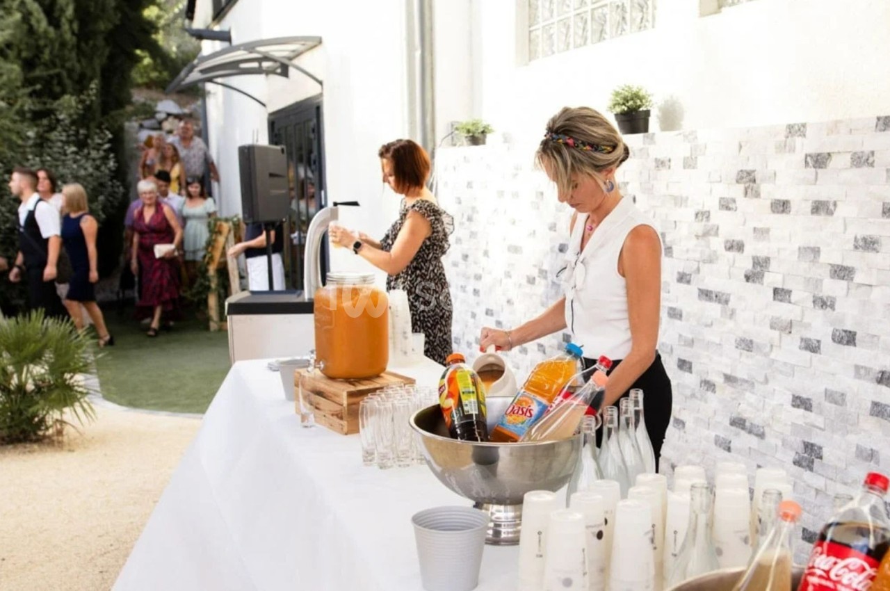 Deux femmes préparent un buffet de boissons en extérieur, avec des invités en arrière-plan près d'un jardin.