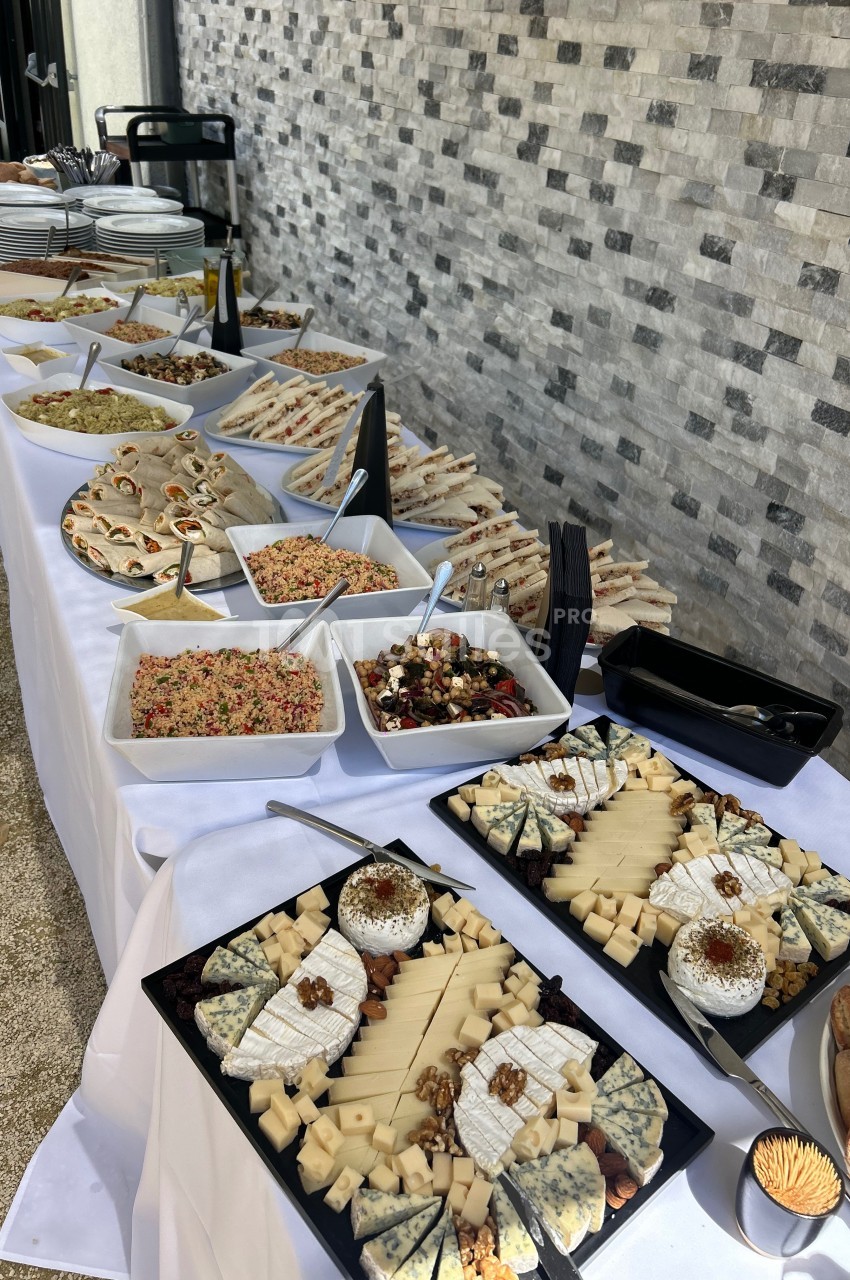 Buffet avec plateaux de fromages, salades variées et sandwichs disposés sur une table blanche en extérieur.