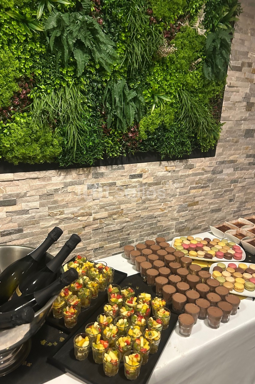 Buffet avec desserts variés, verrines de fruits, macarons et bouteilles de vin, devant un mur végétalisé.