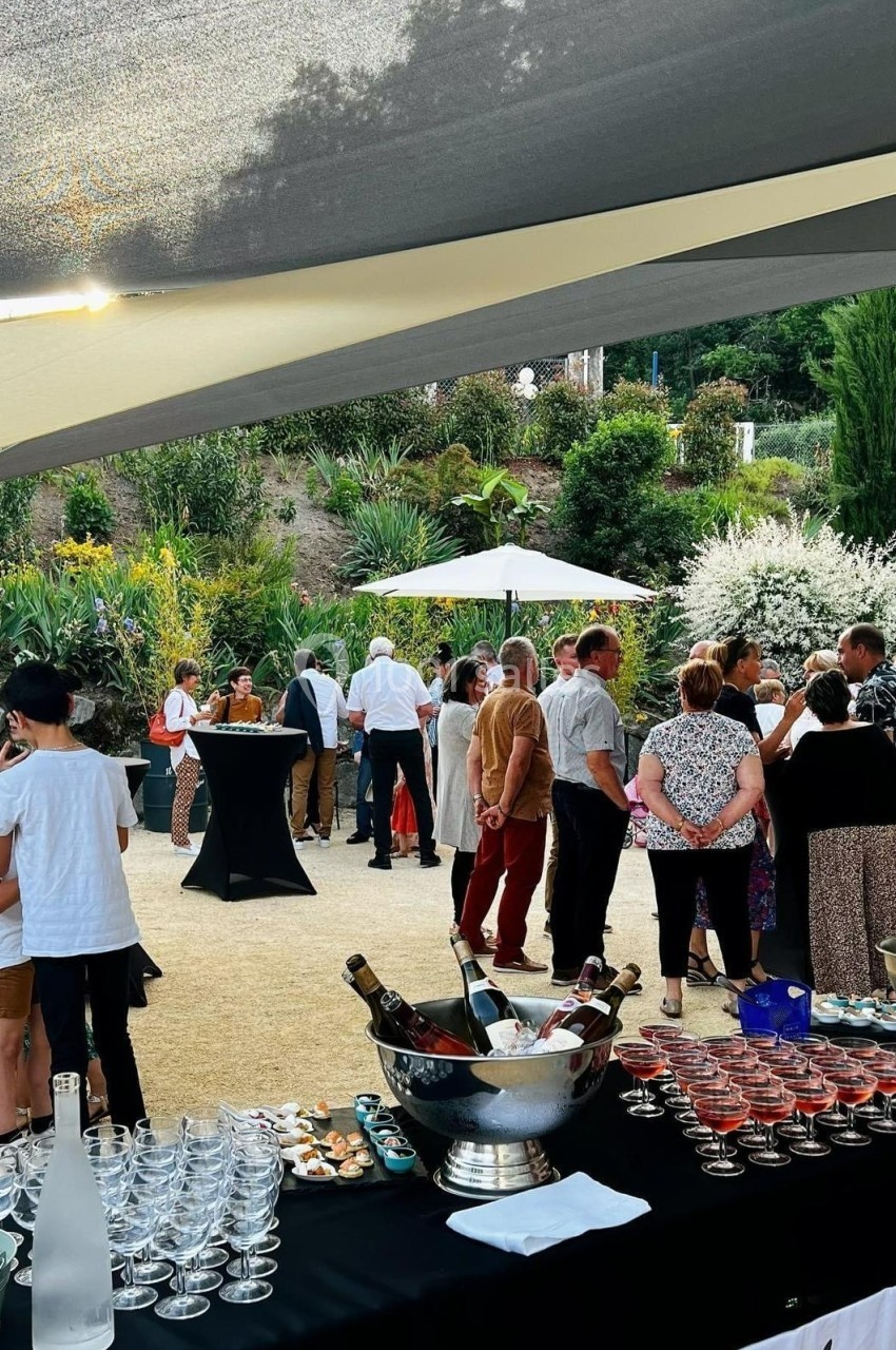 Groupe de personnes discutant lors d'un événement en extérieur, avec tables, boissons et jardin en arrière-plan.