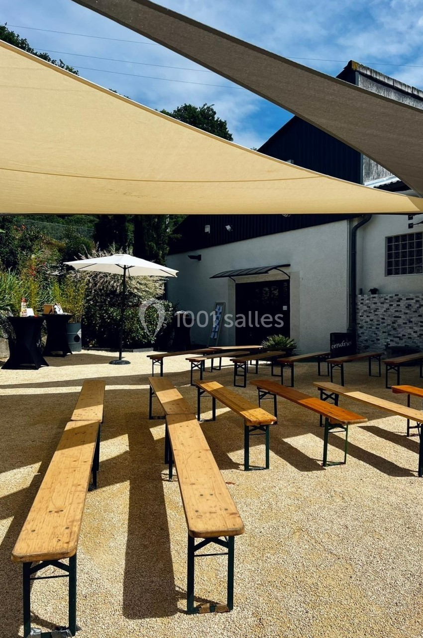 Bancs en bois disposés sur une terrasse en gravier, ombragée par des voiles tendues sous un ciel partiellement dégagé.