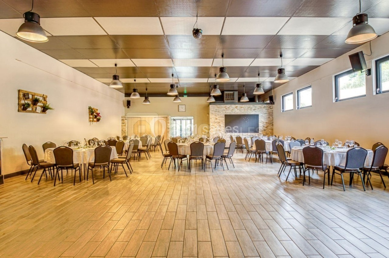 Salle lumineuse aménagée avec des tables rondes dressées, des chaises et un mur en pierre avec une cheminée.
