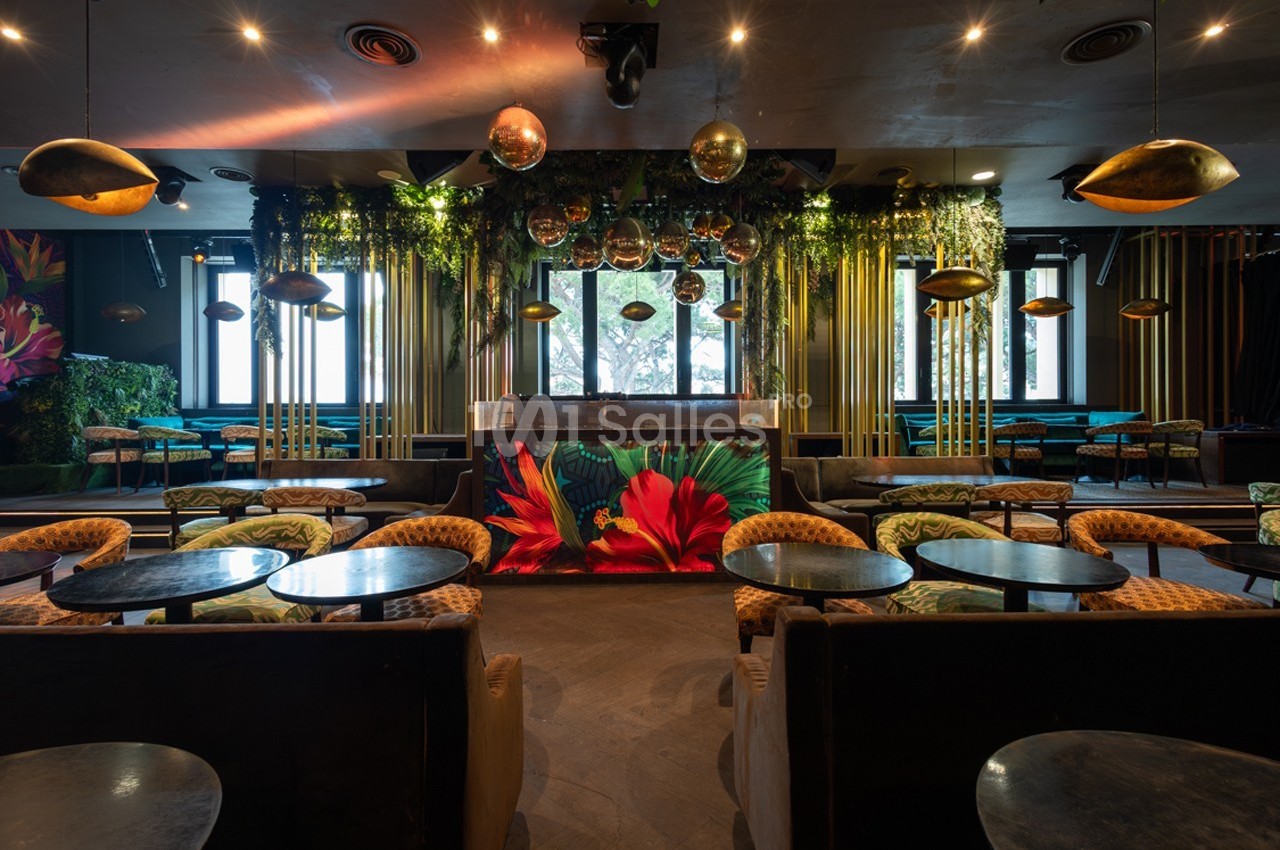 Salle de restaurant décorée de végétation suspendue, luminaires dorés et comptoir orné de motifs floraux colorés.