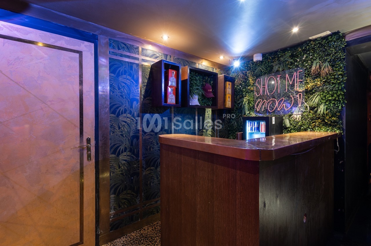 Comptoir en bois dans un bar avec mur végétal, néon ’Shot me now’ et éclairage tamisé.