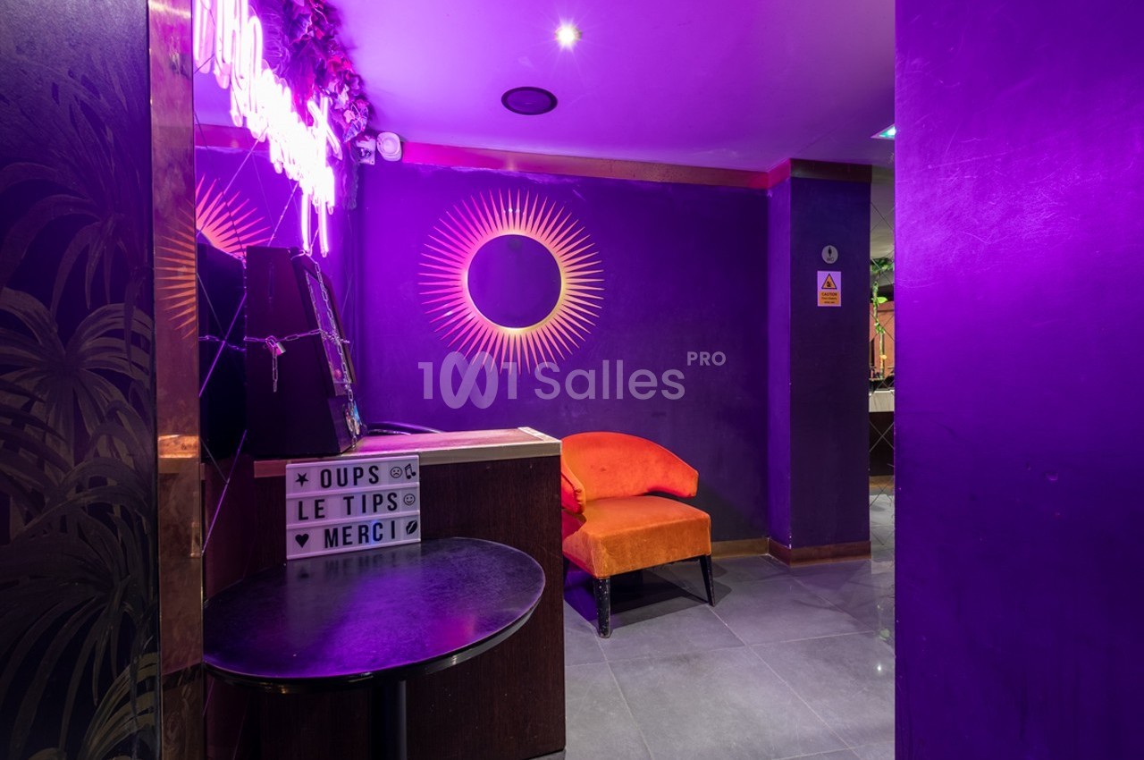 Espace intérieur avec murs violets, chaise orange, néons décoratifs et une boîte lumineuse affichant un message.