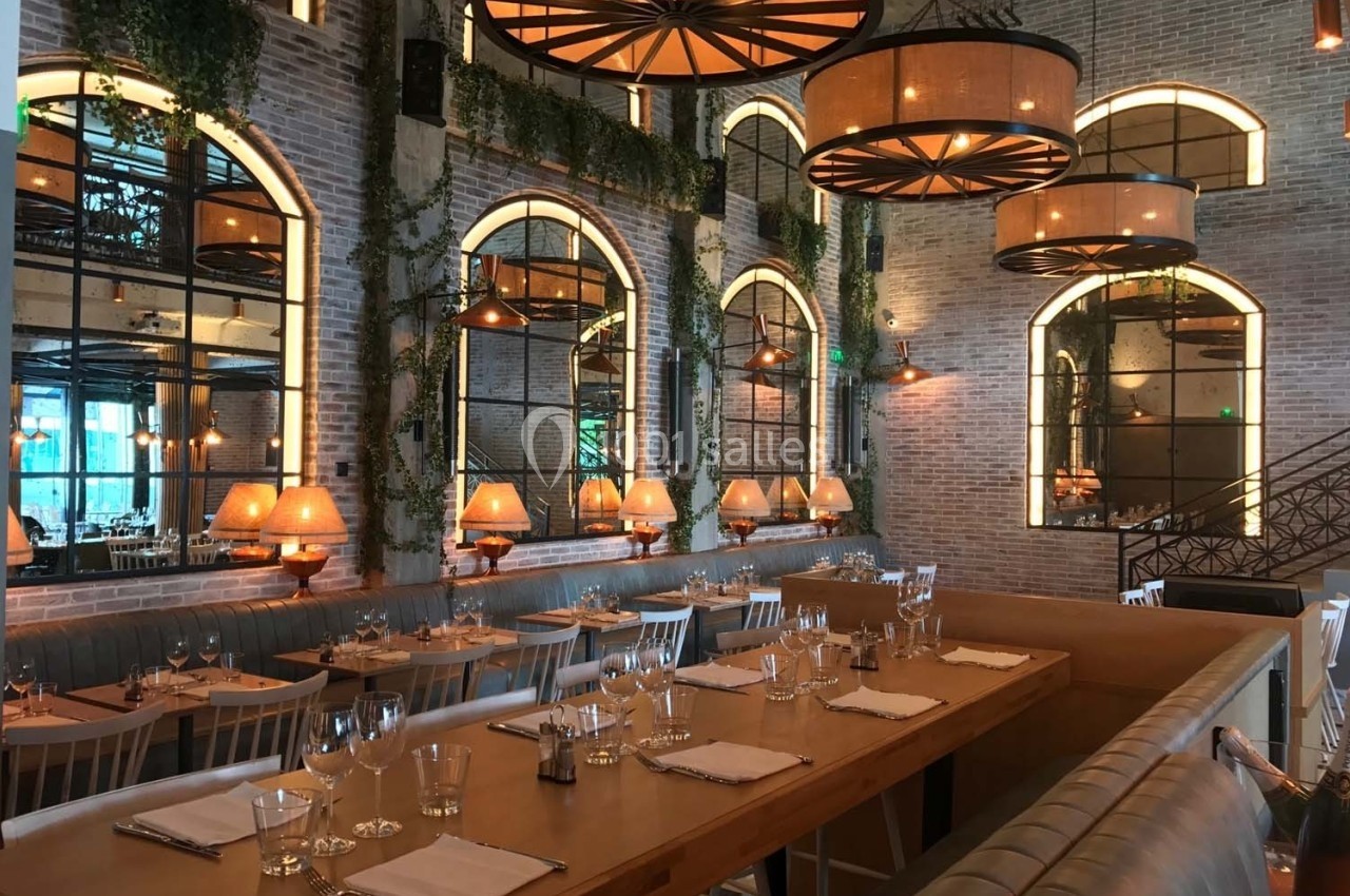 Salle de restaurant lumineuse avec murs en briques, grandes fenêtres, tables dressées et suspensions décoratives.