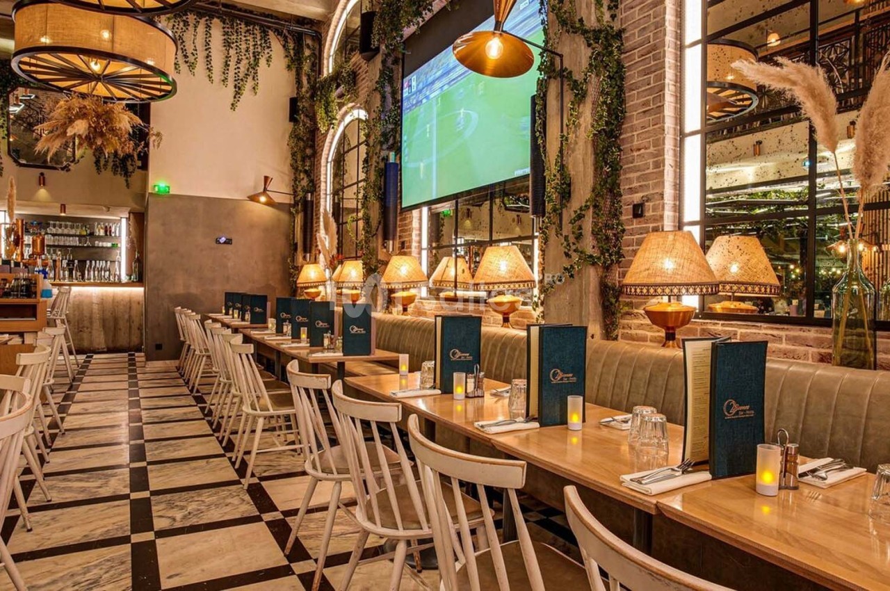 Salle de restaurant chaleureuse avec tables alignées, chaises en bois, éclairage tamisé et décoration végétale.
