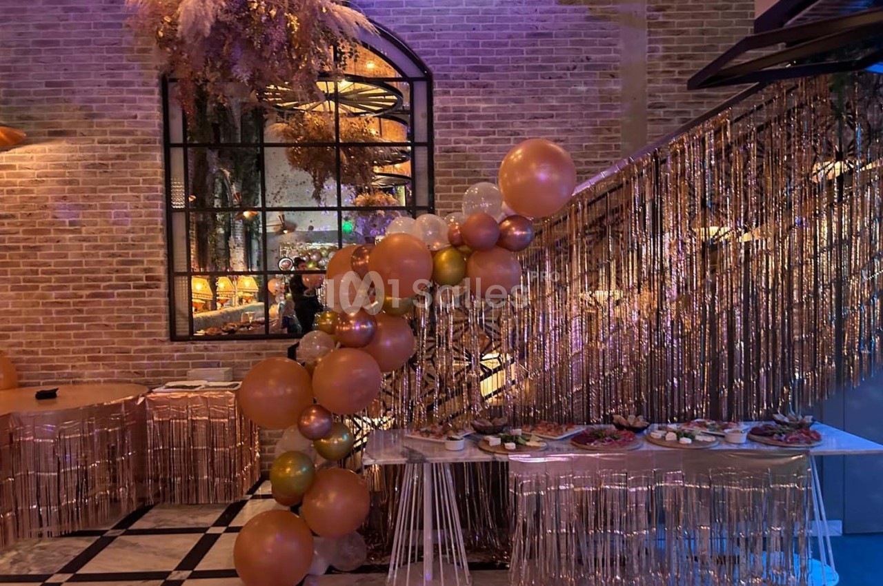 Décoration festive avec ballons dorés et roses, guirlandes scintillantes et table garnie de plats dans une salle en briques.
