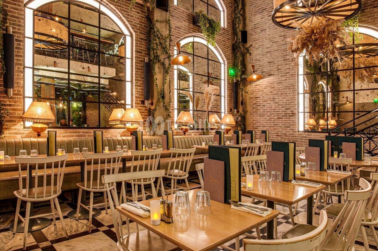 Salle de restaurant chaleureuse avec murs en briques, grandes fenêtres, tables en bois et éclairage tamisé.