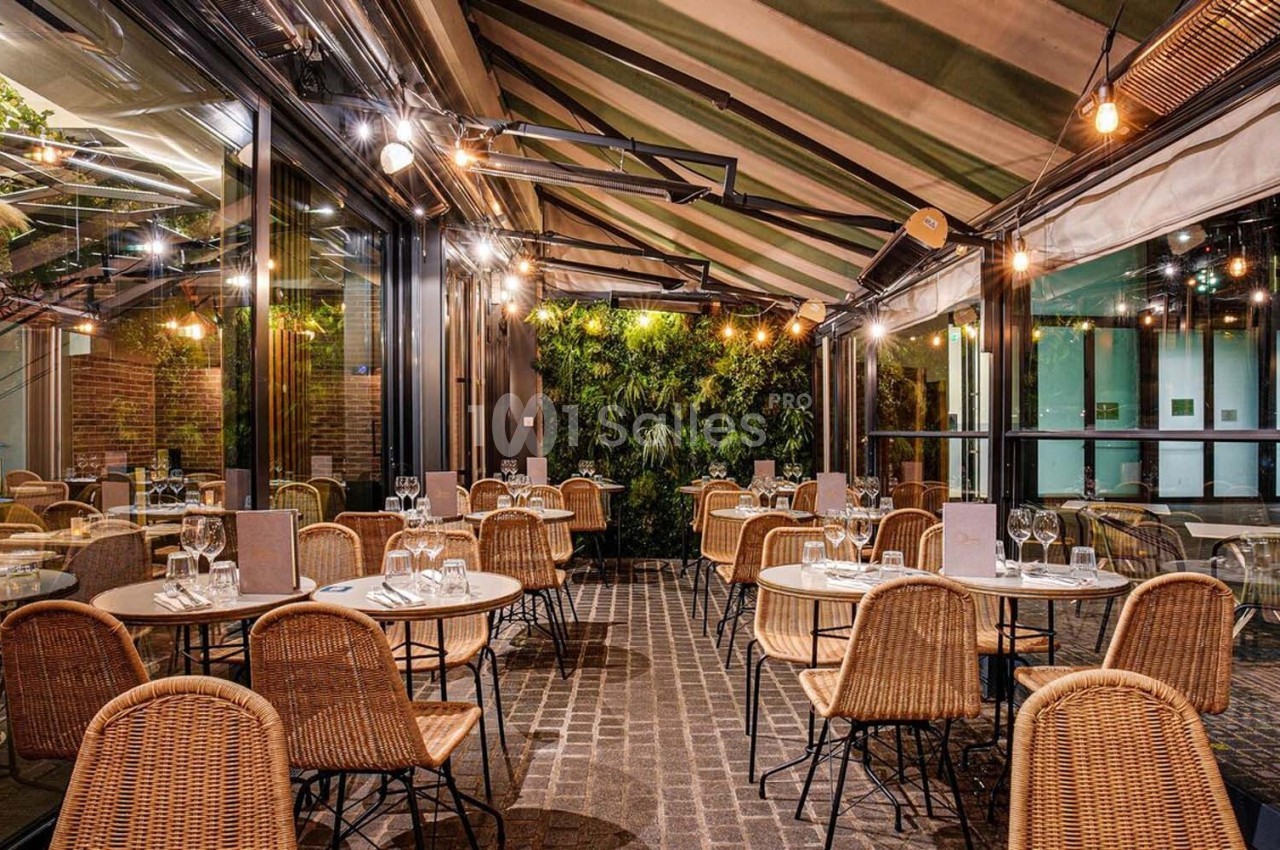 Terrasse couverte avec tables rondes, chaises en rotin, éclairage chaleureux et mur végétal en arrière-plan.