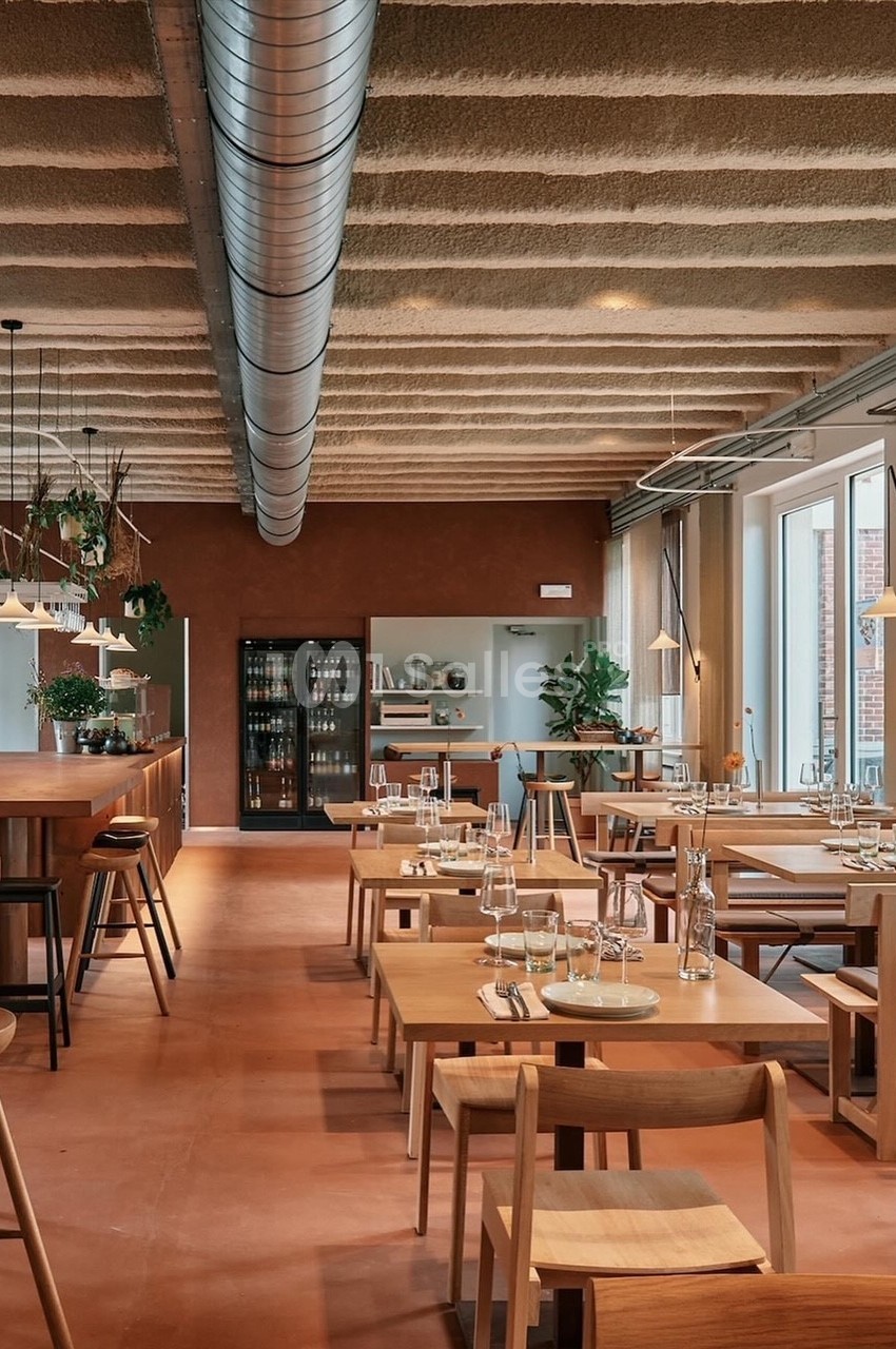 Salle de restaurant moderne avec tables en bois, grandes fenêtres, éclairage suspendu et décoration végétale.